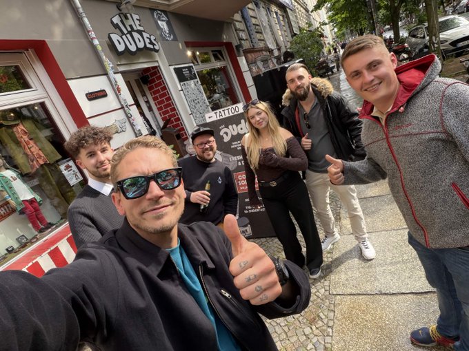 The image features a group of six people posing for a photo outside a tattoo shop called "The Dudes Factory". The individuals are standing close together, with one person giving a thumbs-up gesture as they pose for the picture. In the background, there is a sign that reads "The Dudes Factory" and another sign that says "Merch szn Partnering with @DudesFactory The team behind projects with McBess, BAYC, and other names in art, streetwear, and Web3". This suggests that the group of people might be friends or acquaintances who are interested in tattoos and related activities.
