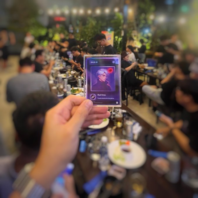 A hand holds up a 'KAZE' trading card featuring an anime-style character at a crowded restaurant gathering, with plates and bottles visible on the table in the background.