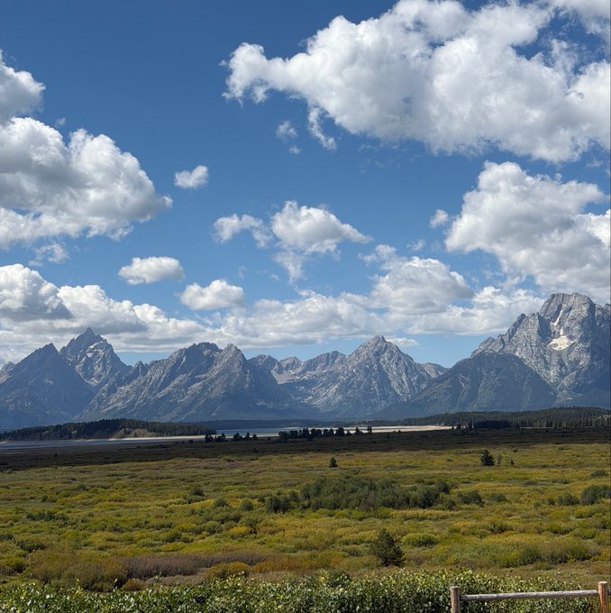 A scenic landscape view of the Grand Teton mountain range with snow-capped peaks rising behind a lush green valley under a bright blue sky filled with scattered white clouds.