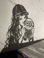 A black and white illustration depicts a person wearing a cap and sunglasses holding a circular object with a face design, positioned against a dark background.