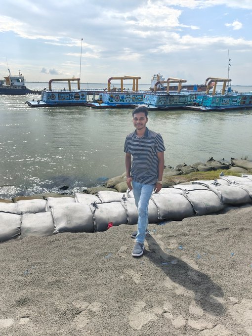 A young man in a striped shirt and jeans stands on a sandy shoreline near large white sandbags, with a large blue and white ship docked in the water behind him.
