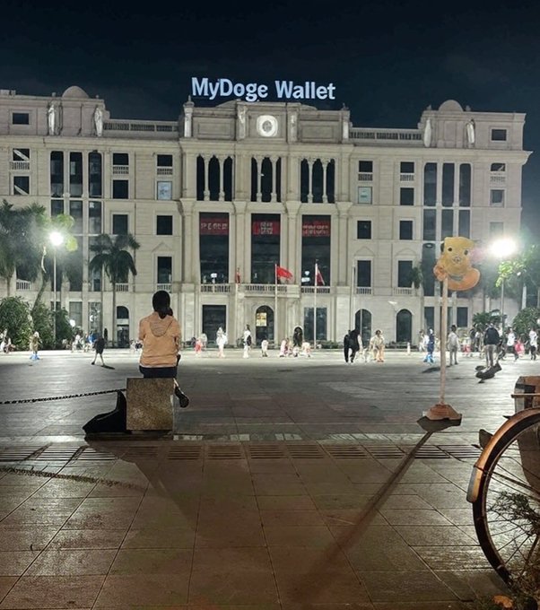 The image shows a person sitting on a bench outside of a large building at night. There are several other people walking around the area as well. The scene also includes a bicycle parked nearby, and a sign that reads "MyDOGE Wallet". This suggests that the location might be related to cryptocurrency or digital currency in some way.