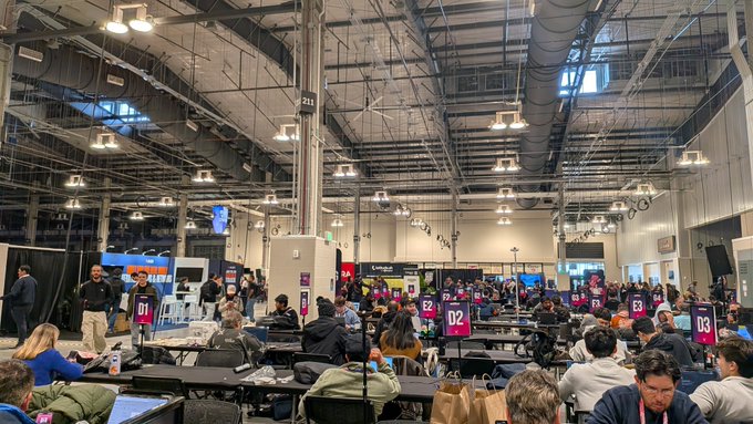 A crowded convention hall or tournament venue with attendees seated at long tables, featuring numbered signs like D1, D2, and D3, under an industrial ceiling with exposed ductwork.