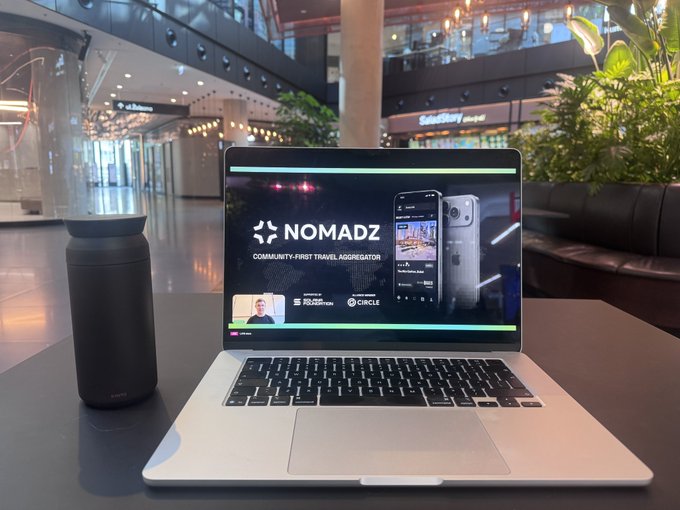 An open laptop on a cafe table displays a presentation for 'NOMADZ - COMMUNITY-FIRST TRAVEL AGGREGATOR,' alongside a dark travel mug in a modern building setting.