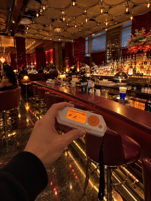 A hand holds a Flipper Zero device in the foreground of an upscale, dimly lit bar with leather stools, warm lighting, and rows of liquor bottles behind the counter—likely capturing a moment of hacking or RFID/NFC exploration at a fancy venue, prompting the awed reaction "Holy shit."
