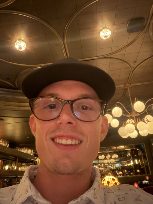 A selfie of a smiling young man wearing black-rimmed glasses and a black cap, with ornate chandeliers visible on the ceiling behind him.