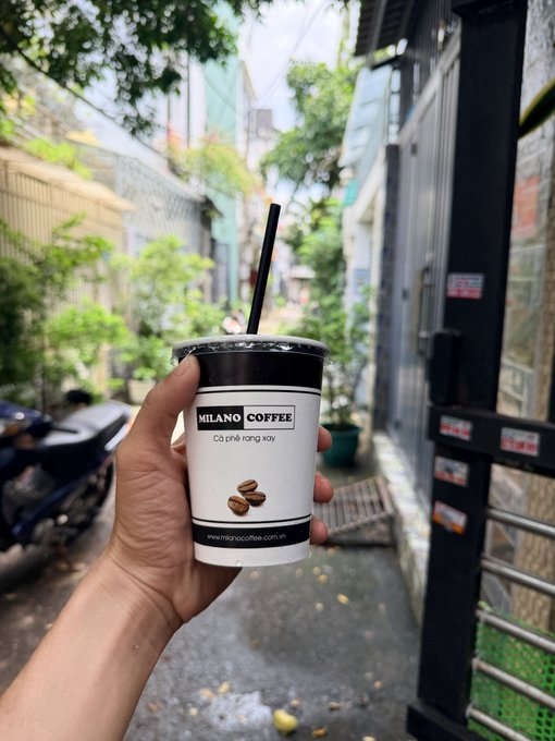A hand holds a cup of Milano Coffee labeled 'Cà phê rang xay' in a narrow Vietnamese alleyway.