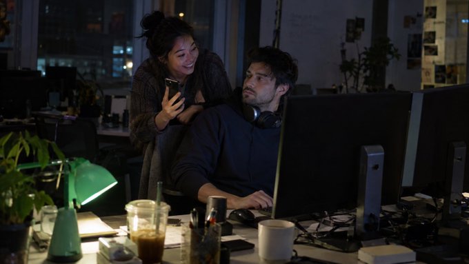 Two people in a dimly lit office at night, with a woman smiling and holding a phone while looking over the shoulder of a seated man wearing headphones at a desk with monitors.