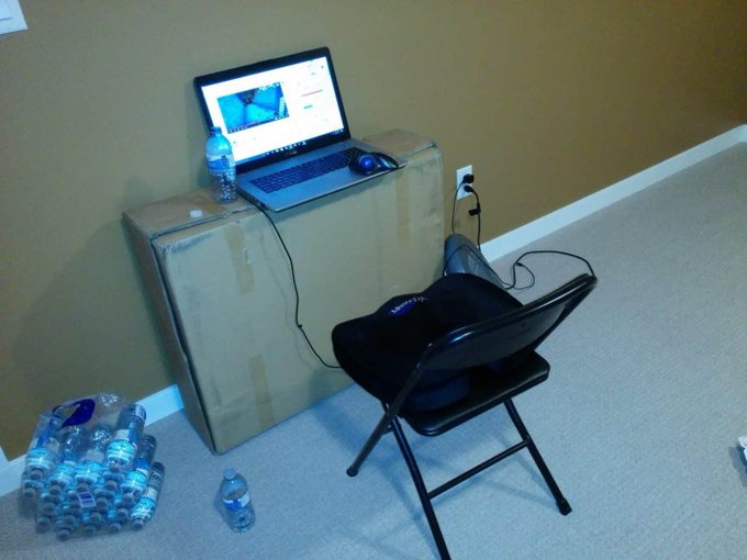 An improvised workstation features a laptop set up on top of cardboard boxes, accompanied by a folding chair and a large stack of bottled water, suggesting someone is prepared for an extended stay. The makeshift setup humorously evokes the early pandemic work-from-home era referenced in the tweet about things "feeling like 2021 again."