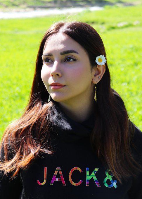 A young woman with long brown hair and a daisy hair clip poses in a field, wearing a black hoodie that says 'JACK&'.