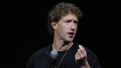 A man with curly brown hair speaks into a microphone while gesturing with his hand against a dark background.