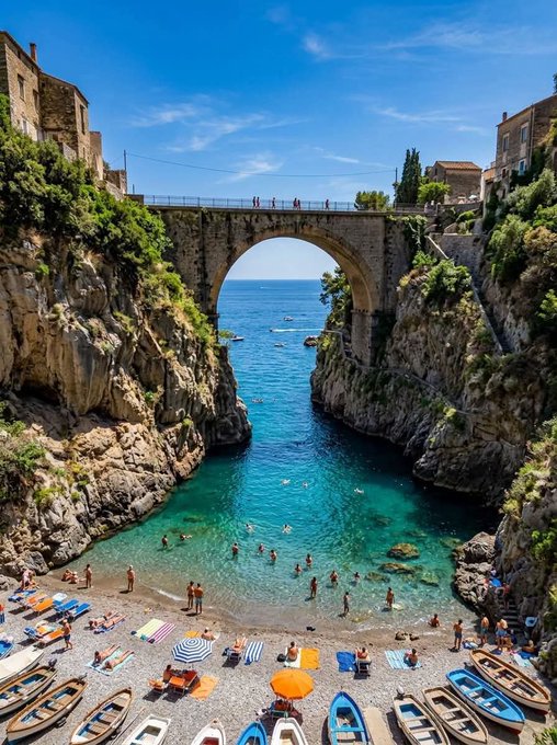 A stone arch bridge spans a turquoise coastal cove where sunbathers relax on a rocky beach below, with small boats moored along the shore—evoking the imagery of bridges connecting separate shores, much like how the post highlights $RIVER's bridge-free cross-chain movement of satUSD between ecosystems.