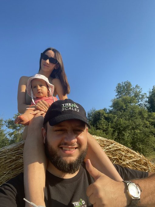 A man wearing a black 'TREND SCALEZ' cap gives a thumbs-up, with a woman in sunglasses and a baby in a pink sun hat sitting on his shoulders against a blue sky and hay bales.