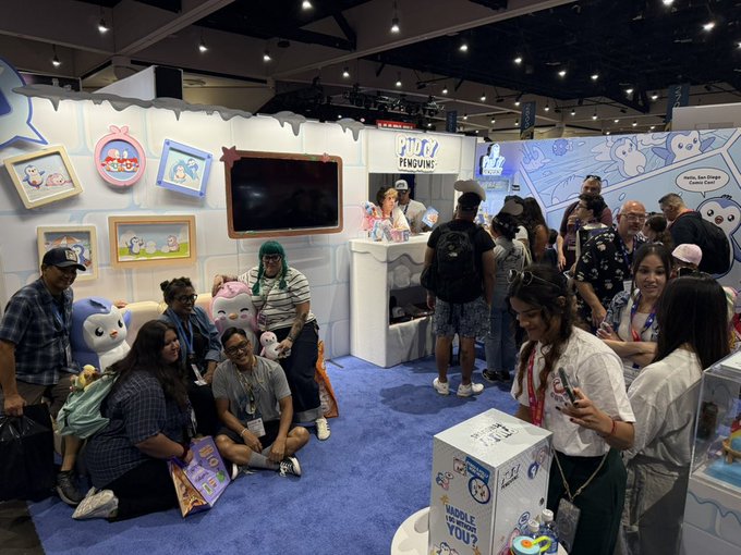 A crowded convention booth for "Tiny Penguins" featuring blue penguin plushies, attendees, and event signage.
