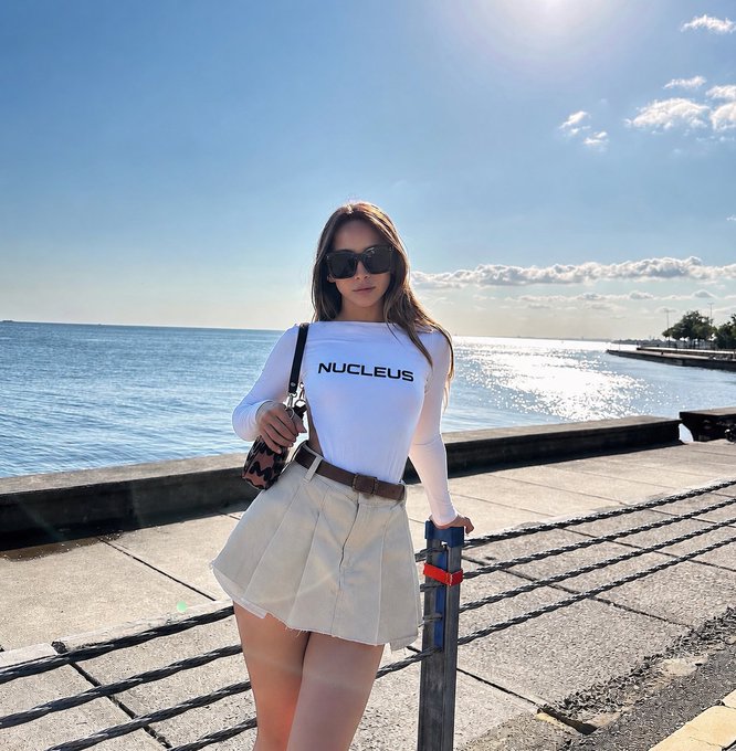 A young woman wearing sunglasses, a white 'NUCLEUS' long-sleeve top, and a beige pleated skirt standing by a waterfront with the ocean in the background.