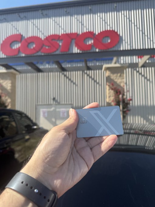 A first-person photo of a hand holding a gray Visa debit/credit card in front of a Costco store entrance, with a parked car visible below.
