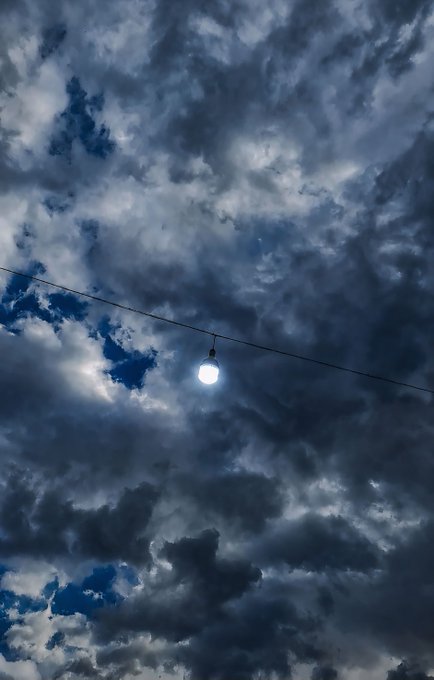 A single illuminated streetlight bulb hangs from a wire against a dramatic backdrop of dark, swirling storm clouds with patches of blue sky visible.