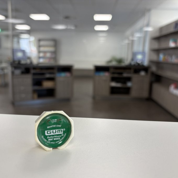 A small octagonal container of GUM brand mint waxes sitting on a white counter with a blurred convenience store or pharmacy background.