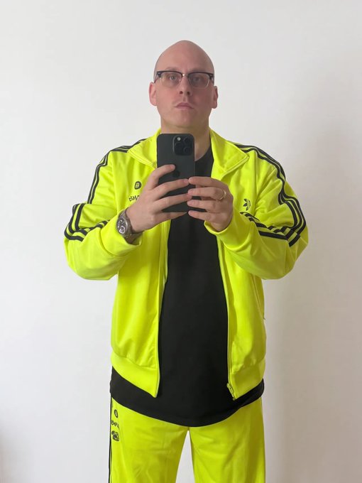 A bald man wearing glasses and a bright neon yellow tracksuit takes a mirror selfie with a black smartphone.