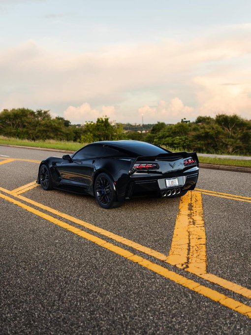 A black Chevrolet Corvette sports car is parked on a road with yellow lines under a cloudy sky at dusk.