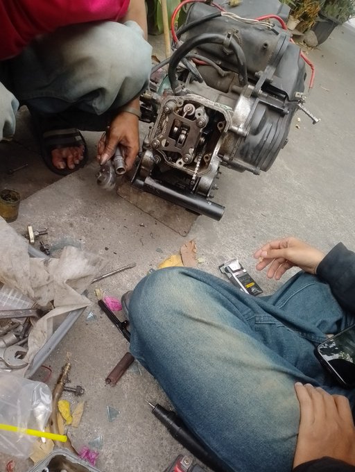 A mechanic works on repairing a motorcycle engine, with tools and parts scattered around, while someone in jeans sits nearby, illustrating a hands-on "kia rehab mesin duluu" (machine rehabilitation) session.
