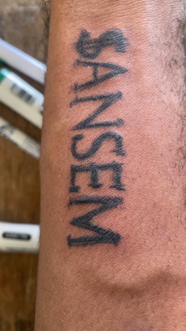 A close-up photo of the word "ANSEM" tattooed in bold black capital letters on a person's forearm, matching the post about the user getting the ticker inked as a tribute to the crypto figure who "saved the trenches."