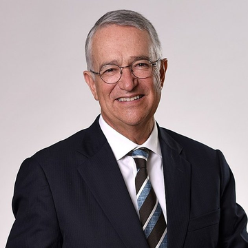 The image shows a smiling older man with gray hair and glasses, wearing a dark suit and a blue striped tie, posing for a professional headshot against a white background. The photo likely represents the Mexican billionaire referenced in the post as the country's third-richest man, who has reportedly allocated 70% of his personal portfolio to Bitcoin.