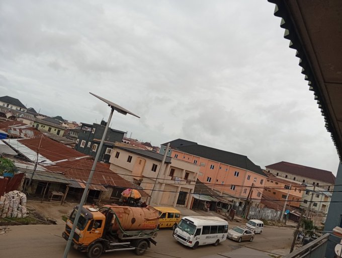 A street view of a town with an orange sewage truck and white mini-buses parked on the road, set against a backdrop of buildings with corrugated metal roofs under a cloudy sky.