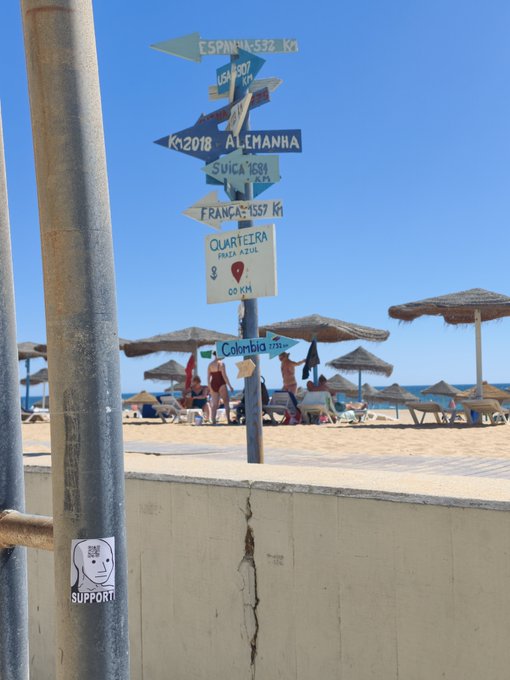 A signpost at a beach points to various locations with their distances, including 'ESPANHA 532 KM', 'RUSSIA' (partially visible), 'ALEMANHA Km 2018', 'SUIÇA 1681', 'FRANÇA 1557 KM', 'QUARTEIRA PRAIA AZUL 0,00 KM', and 'Colombia'. A sticker reading 'GAME OVER! SUPPORT' with a sad face is attached to the pole.