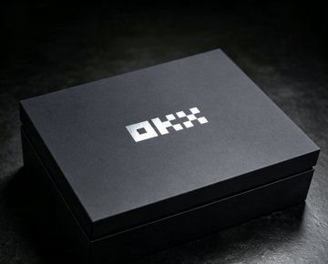 A matte black square box with 'OKX' printed in white lettering on the lid, sitting on a dark surface.