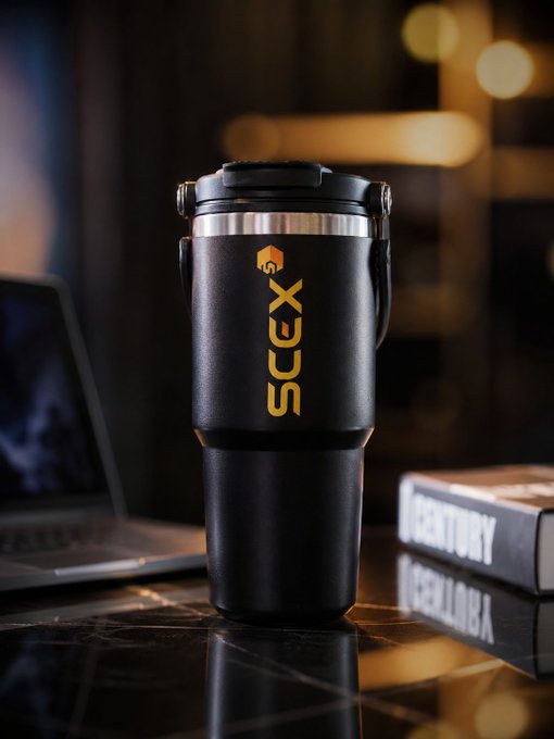A product photo of a black insulated travel mug/tumbler with "SCEX" branding in gold lettering, placed on a dark surface next to a laptop and book.