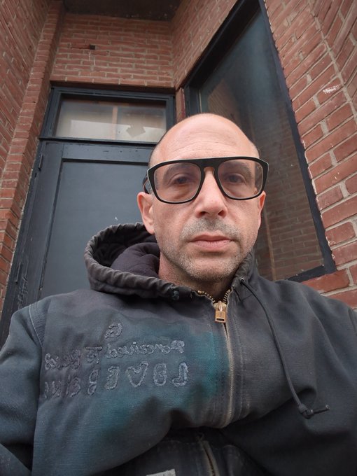 A bald man wearing glasses and a dark hoodie with embossed text stands in front of a brick building with a dark door.