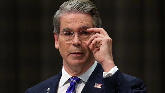 A portrait of Scott Bessent, the U.S. Treasury Secretary, adjusting his glasses while wearing a dark pinstripe suit and blue tie.