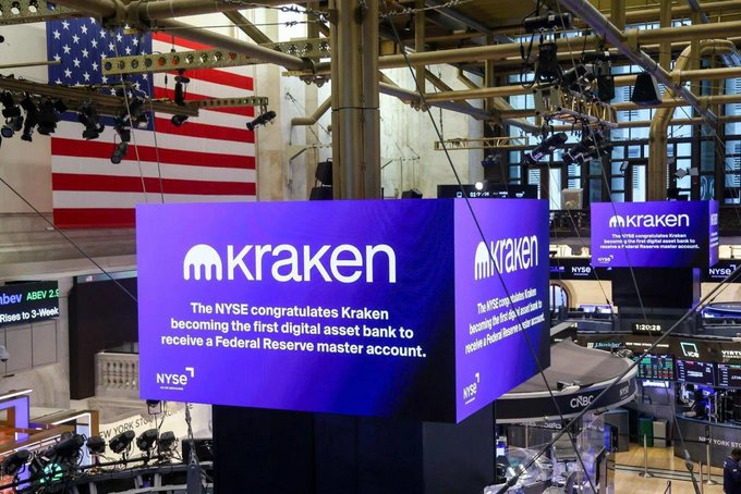 A digital display at the New York Stock Exchange shows the Kraken logo and reads 'The NYSE congratulates Kraken becoming the first digital asset bank to receive a Federal Reserve master account,' with an American flag in the background.
