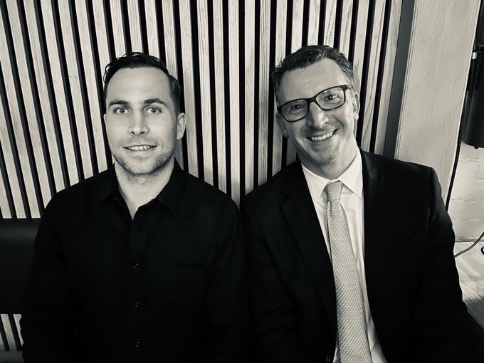 Black-and-white portrait of two men smiling, one younger man in a dark shirt and an older man in a suit with glasses, posed together against a slatted wood background.