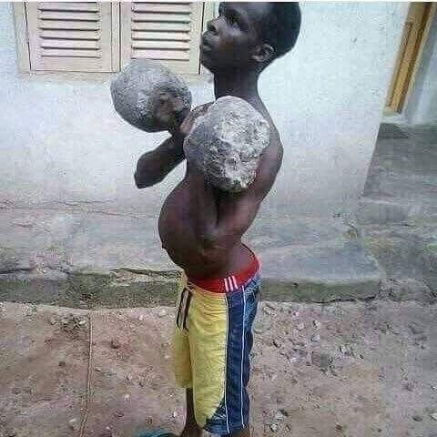 A young boy stands outdoors shirtless, posing as a boxer with makeshift gloves made of stones or concrete.