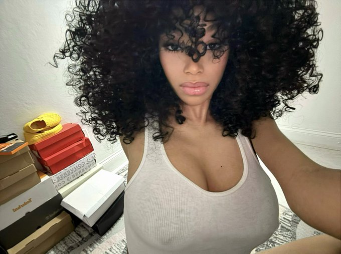 A woman with voluminous curly hair takes a selfie wearing a ribbed tank top, posing in front of several designer shoe boxes including Yeezys and what appears to be Christian Louboutin boxes, perfectly embodying the caption "The new owner of ur wallet" with her confident, sultry gaze.