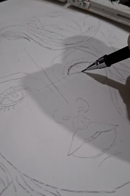 A close-up of an artist's hand using a mechanical pencil to sketch a detailed portrait of a face with large eyes on white paper, with a handwritten signature visible.