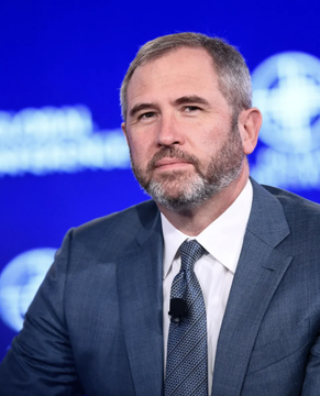 Brad Garlinghouse, CEO of Ripple, appears on stage at what appears to be the Milken Institute Global Conference, shown in a dark suit and tie with a lapel microphone, as he discusses criticism of Michael Saylor's Bitcoin acquisition strategy.