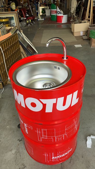 A red Motul oil barrel repurposed into a sink with a stainless steel basin and chrome faucet, displayed in what appears to be a warehouse or antique store setting.