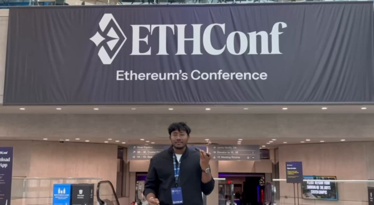 The image features a man standing next to a large banner that says "ETHCONF" written in white letters. The man appears to be wearing a name tag, possibly indicating his role or position at an event related to cryptocurrency. He is giving a thumbs up gesture towards the camera and seems to be enjoying himself.