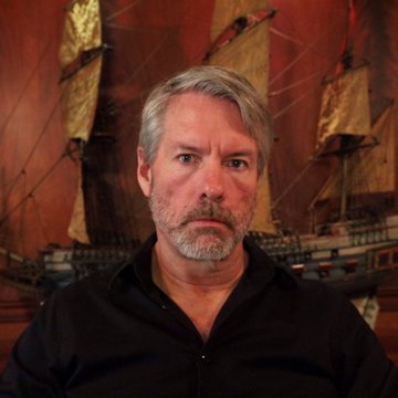A gray-haired, bearded man in a black polo shirt stands in front of a model ship, his serious expression matching Michael Saylor's cryptic "Even more orange" post that preceded Strategy's 950 BTC purchase announcement.