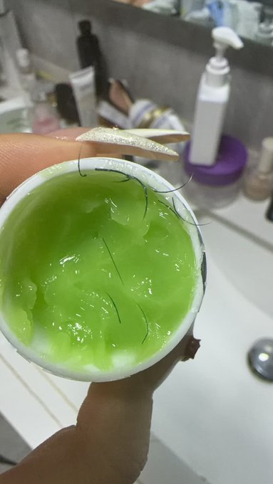 A hand holds a white container filled with green gel used for eyelash extension removal, with several stray lashes stuck in it, while holding a tool to pluck more out. The image illustrates the frustrating process of removing low-quality false lashes from a new salon, contrasting them with the better lashes previously obtained from the original shop.