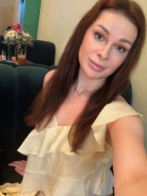 Selfie of a woman with long auburn hair wearing a pale yellow off-shoulder ruffled top, sitting in what appears to be a restaurant with flowers in the background.
