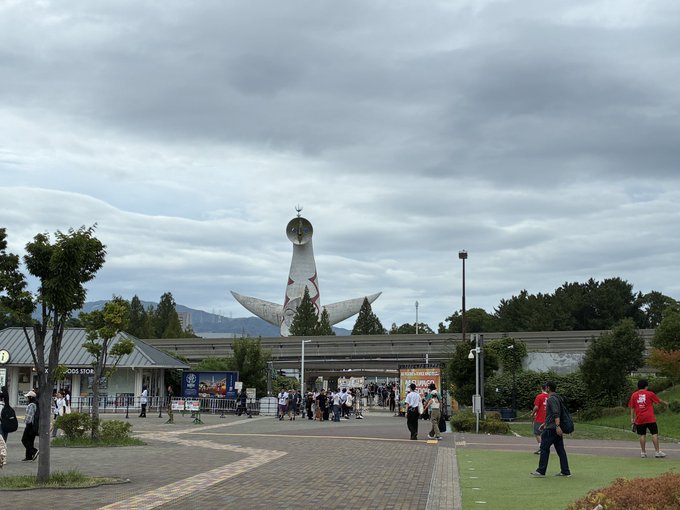 A view of the Tower of the Sun at Expo '70 Commemorative Park in Osaka, with crowds of people gathered near the entrance under a cloudy sky.