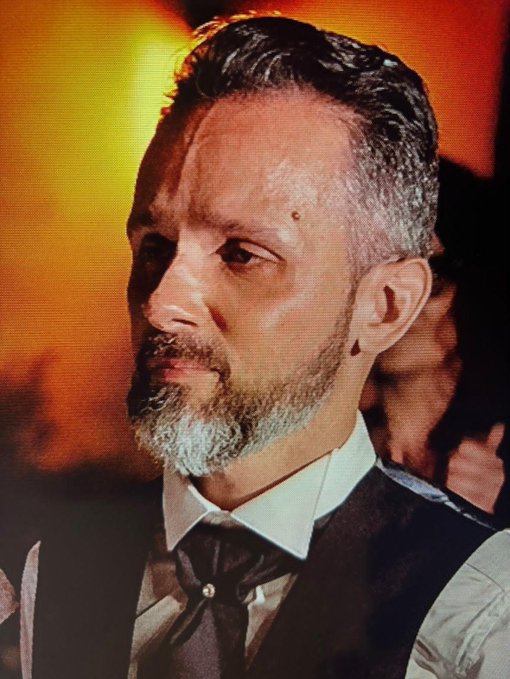 A portrait of a man with short graying hair and a beard, wearing a formal vest with a cravat, set against a warm orange-lit background.