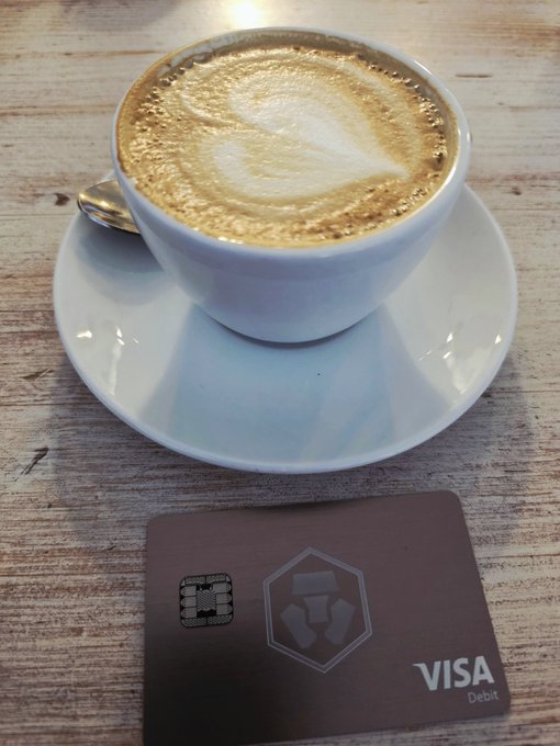 A cappuccino with latte art served in a white cup on a saucer, placed next to a Visa debit card with a crypto wallet logo on a wooden table.