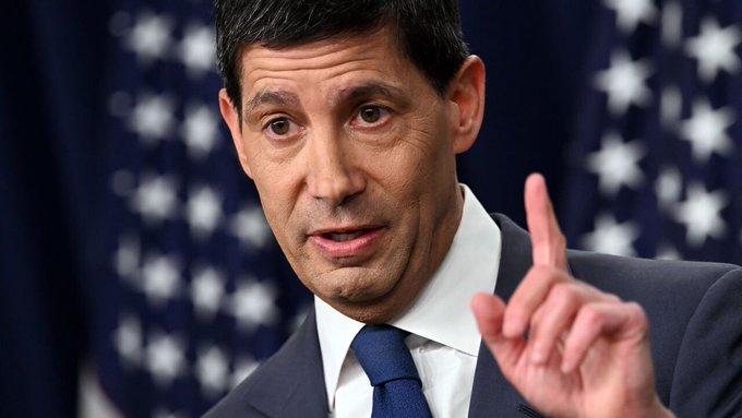 A man in a dark suit and blue tie is pointing his finger upward while speaking, with an American flag visible in the blurred background.