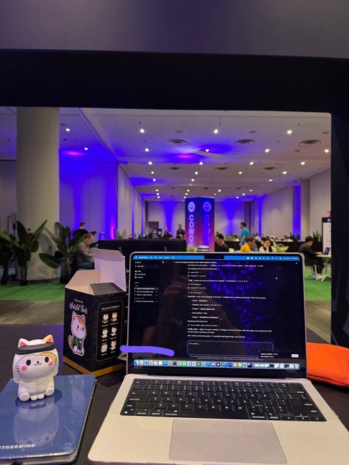 The image shows an open laptop computer sitting on a desk with a mouse next to it. The laptop screen displays a webpage that appears to be related to cryptocurrency or blockchain technology. A small figurine of a cat is also present on the desk, adding a personal touch to the workspace. In the background, there are several people gathered in what seems to be an office setting, possibly engaged in discussions about the content displayed on the laptop screen.