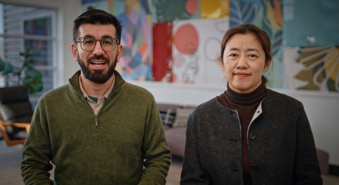 A bearded man in a green pullover and a woman in a dark jacket smile warmly at the camera in a brightly lit room with colorful artwork behind them. Their confident, content expressions reflect the post's message that perseverance leads to clarity and progress after a long journey.